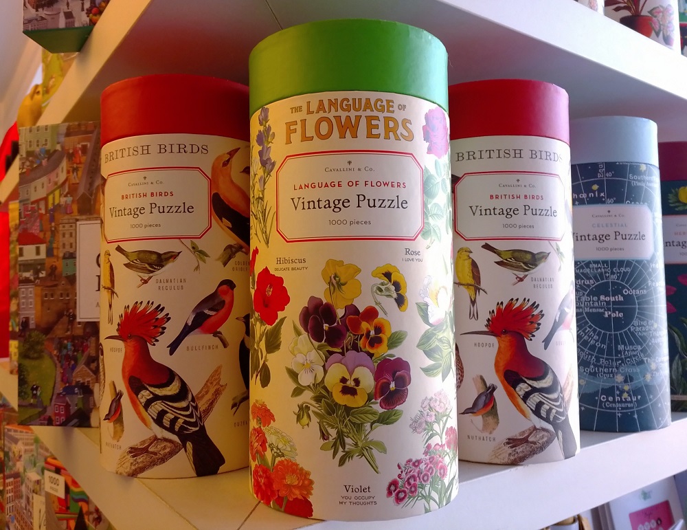 Auf dem Regal stehen mehrere Dosen der Vintage Puzzles, frontal zu sehen Language of Flowers, rechts und links davon stehen jeweils eine Dose mit Motiv British Birds
